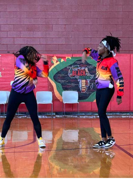 Two dancers perform in a gym in front of a Black History Month banner.